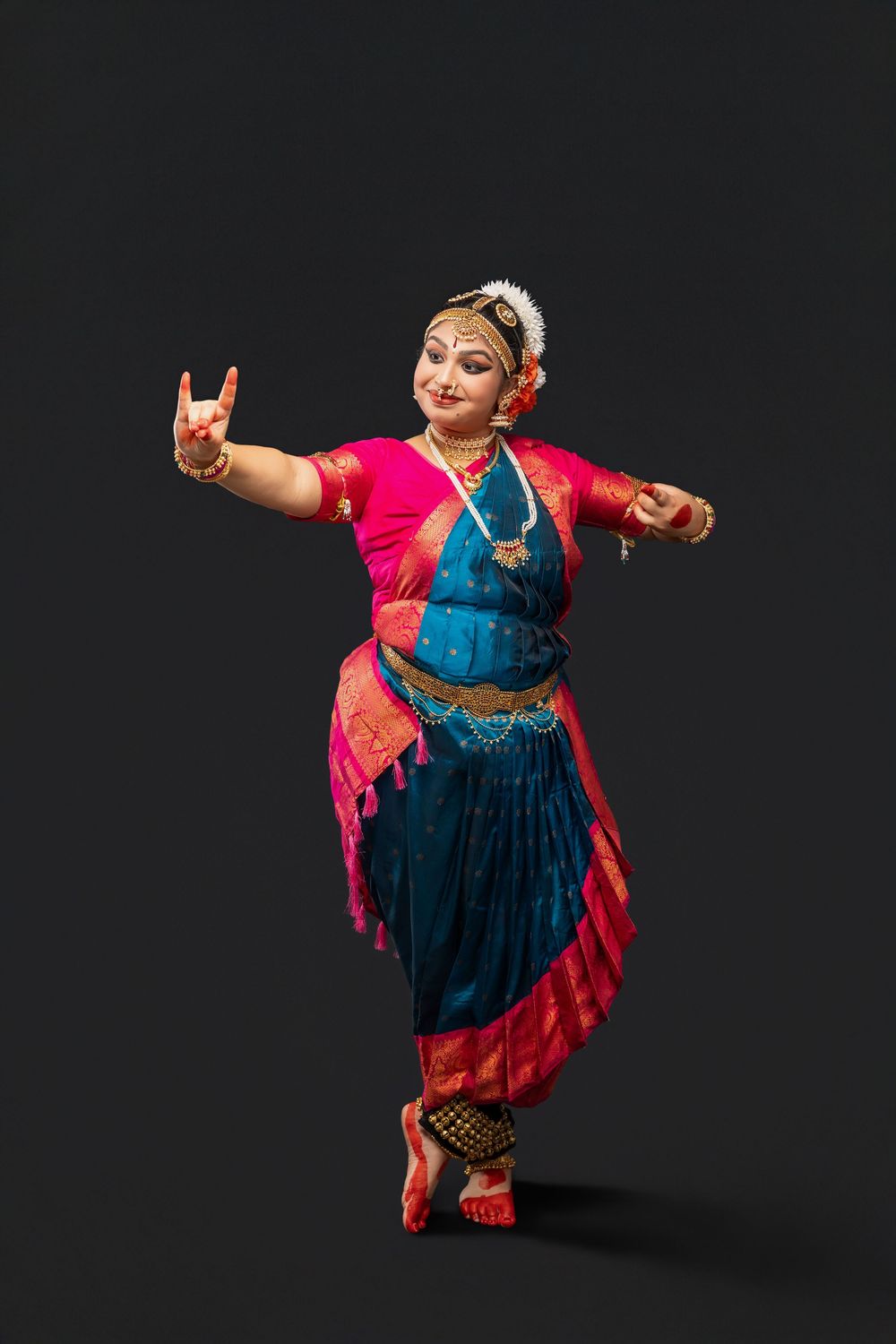 Classical Indian dancer performs in traditional Bharatanatyam costume with vibrant red and blue colors against dark background.