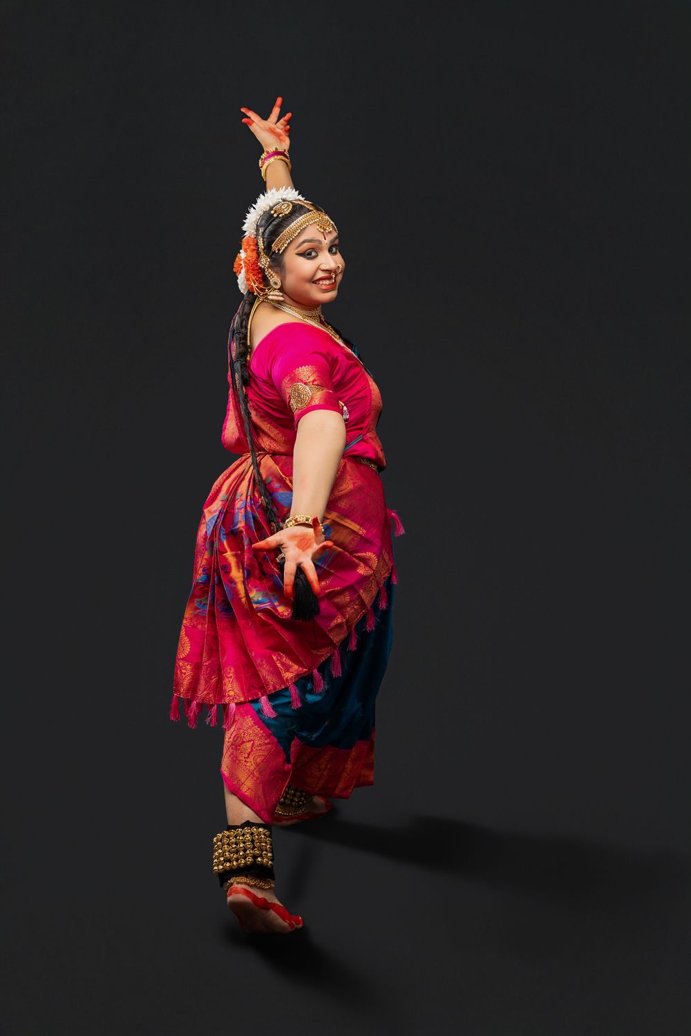 Classical Indian dancer in traditional red and purple sari performs graceful pose against dark background.