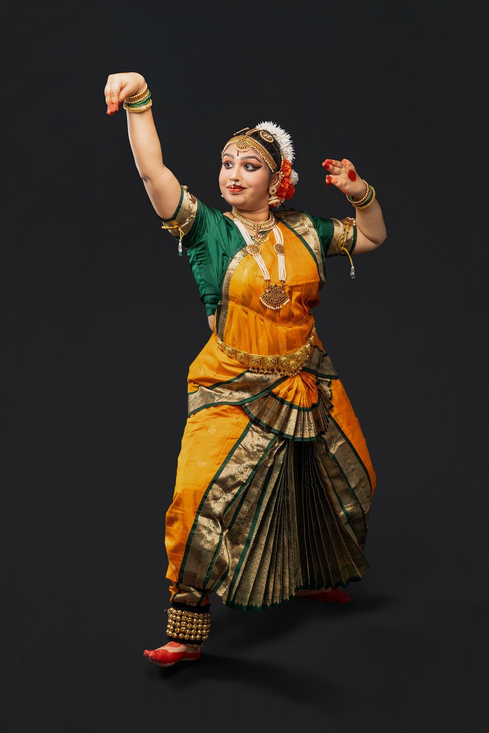 Classical Indian dancer in orange and green traditional costume performs expressive Bharatanatyam dance poses against black backdrop.