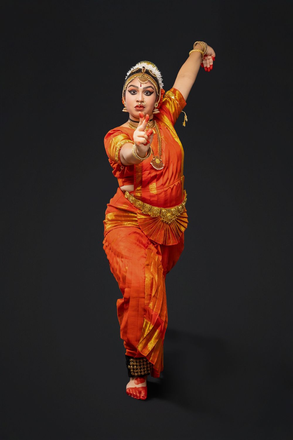 Traditional bharatanatyam dancer in vibrant orange sari performs expressive dance pose on black backdrop.
