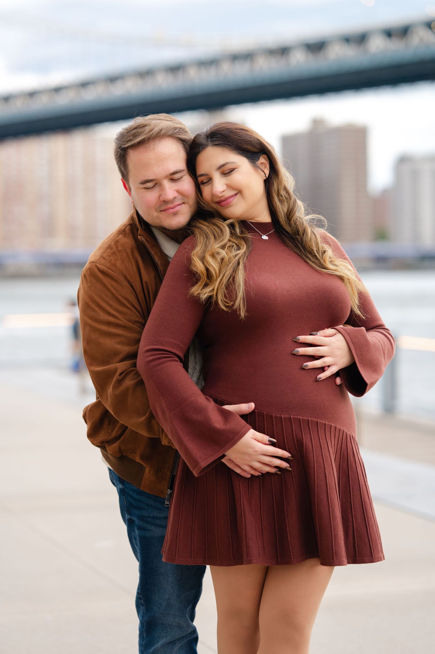A couple poses intimately with their eyes closed for maternity photos in DUMBO in front of the Brooklyn Bridge.