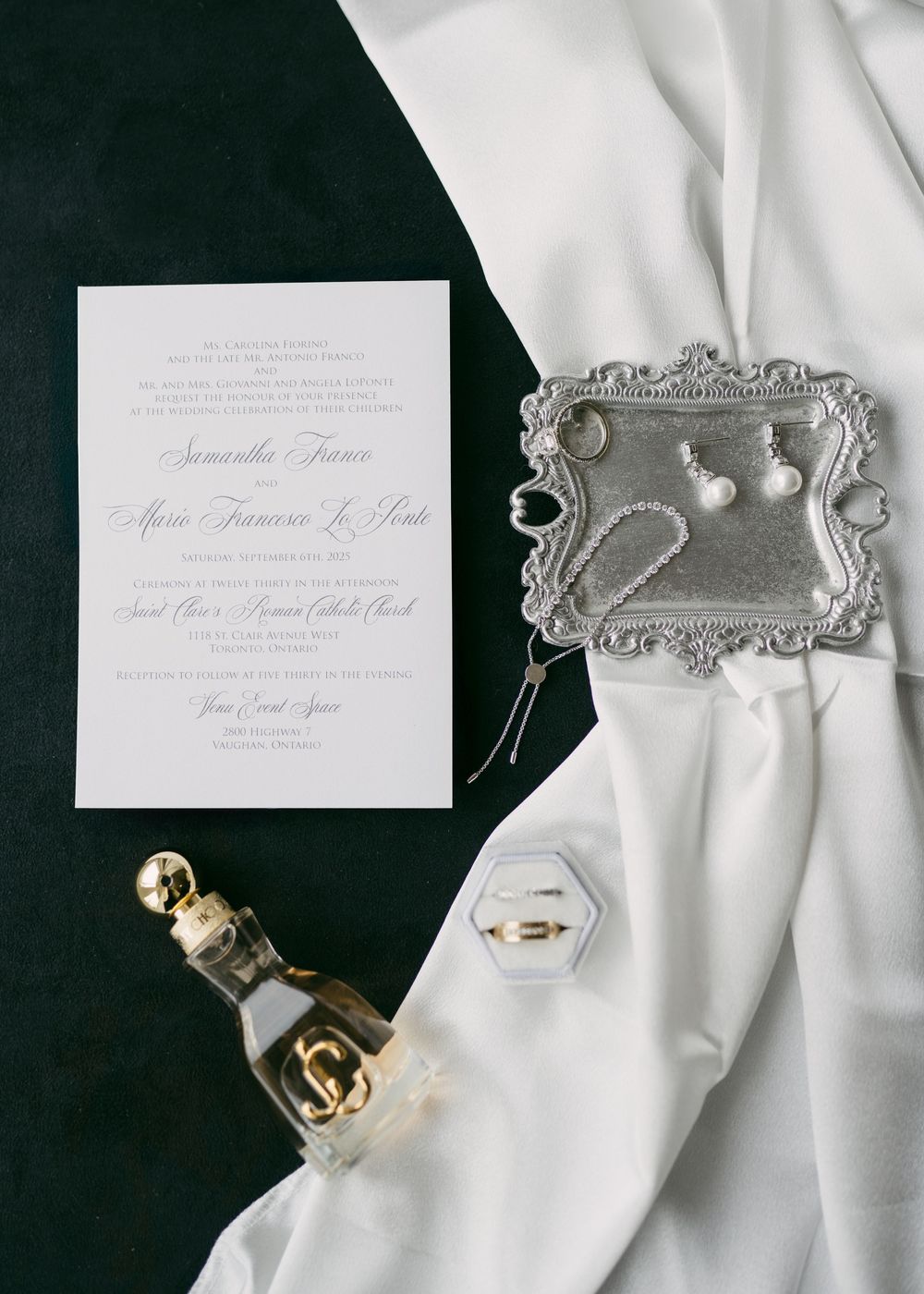 Elegant wedding invitation suite with white stationery, perfume bottle, and jewelry on black background.