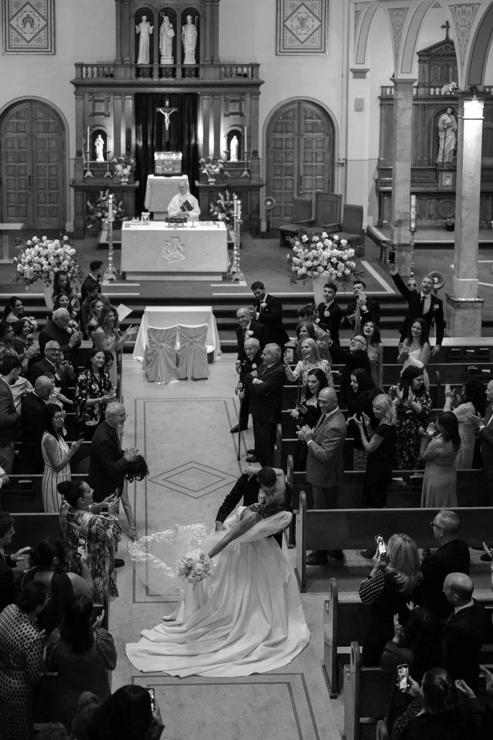 A traditional Catholic church wedding ceremony with people gathered in pews and white floral decorations at the altar.