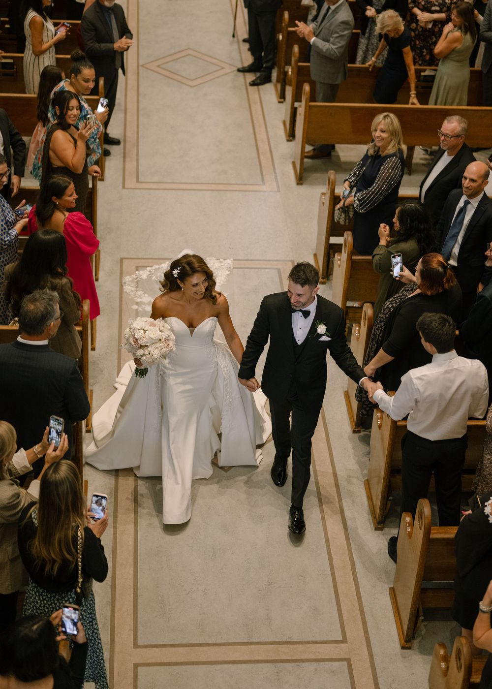 Wedding ceremony procession in a church with guests seated in pews and a white aisle runner.