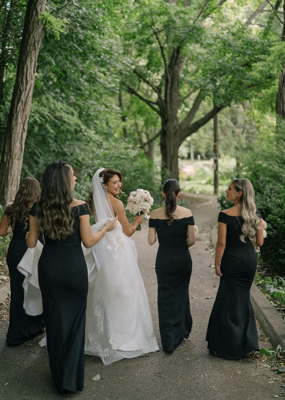 Bridesmaids in black dresses walking with bride in white gown through a tree-lined path.
