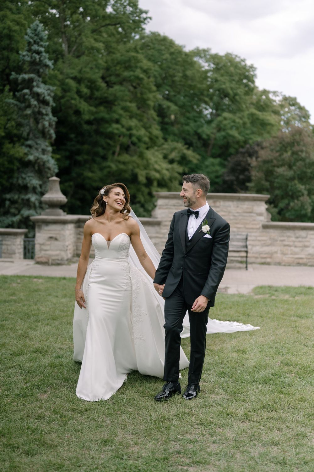 Newlyweds share playful moments on grassy lawn near stone architecture.