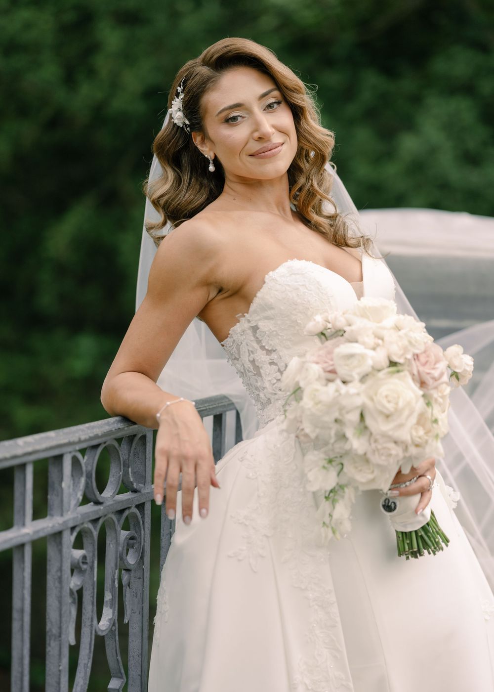 Bride in a white lace wedding dress holds a white floral bouquet while posing on an outdoor railing.