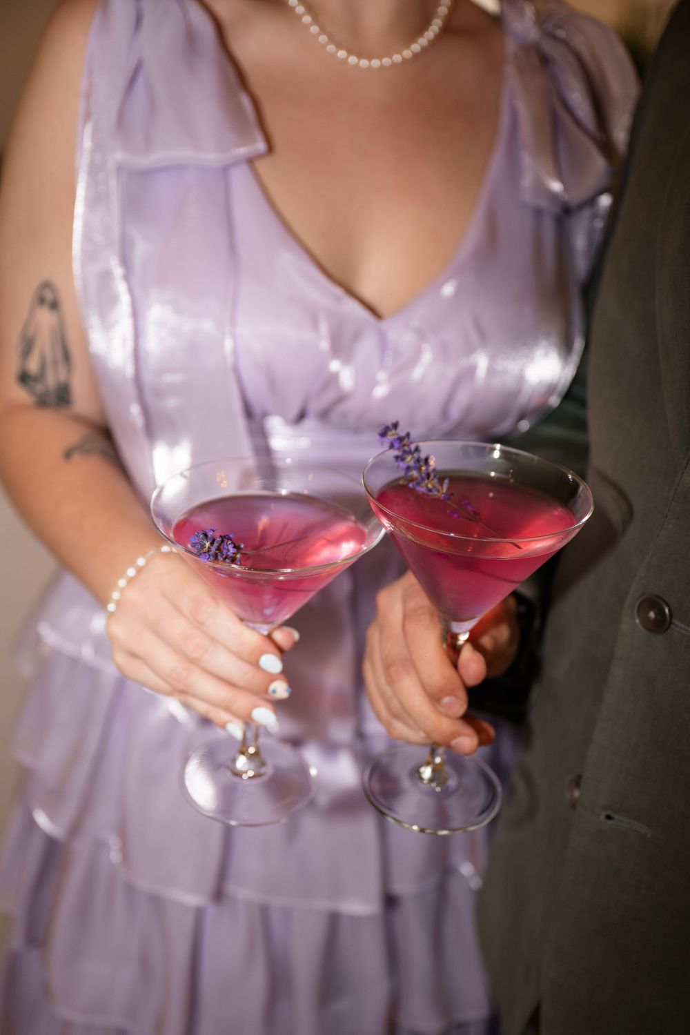 Someone in a lilac dress holds two martini glasses filled with pink cocktails at an elegant event.