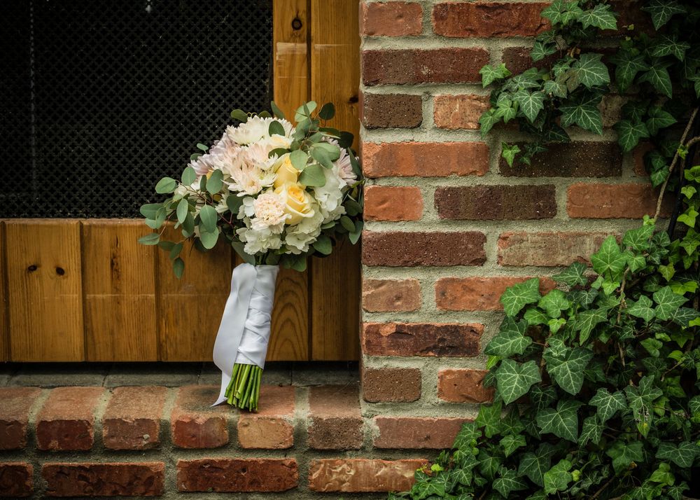 White and green bridal bouquet with roses and eucalyptus leaning against a rustic brick wall.