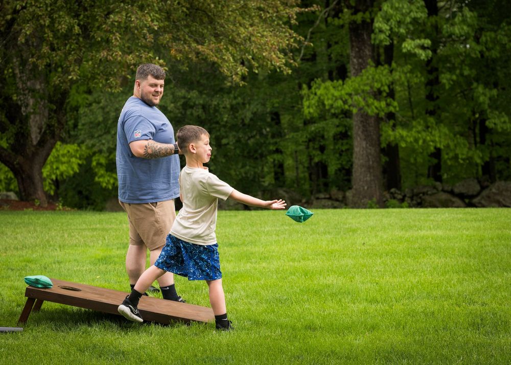 Father and son enjoy outdoor cornhole game on green lawn surrounded by trees at family event.