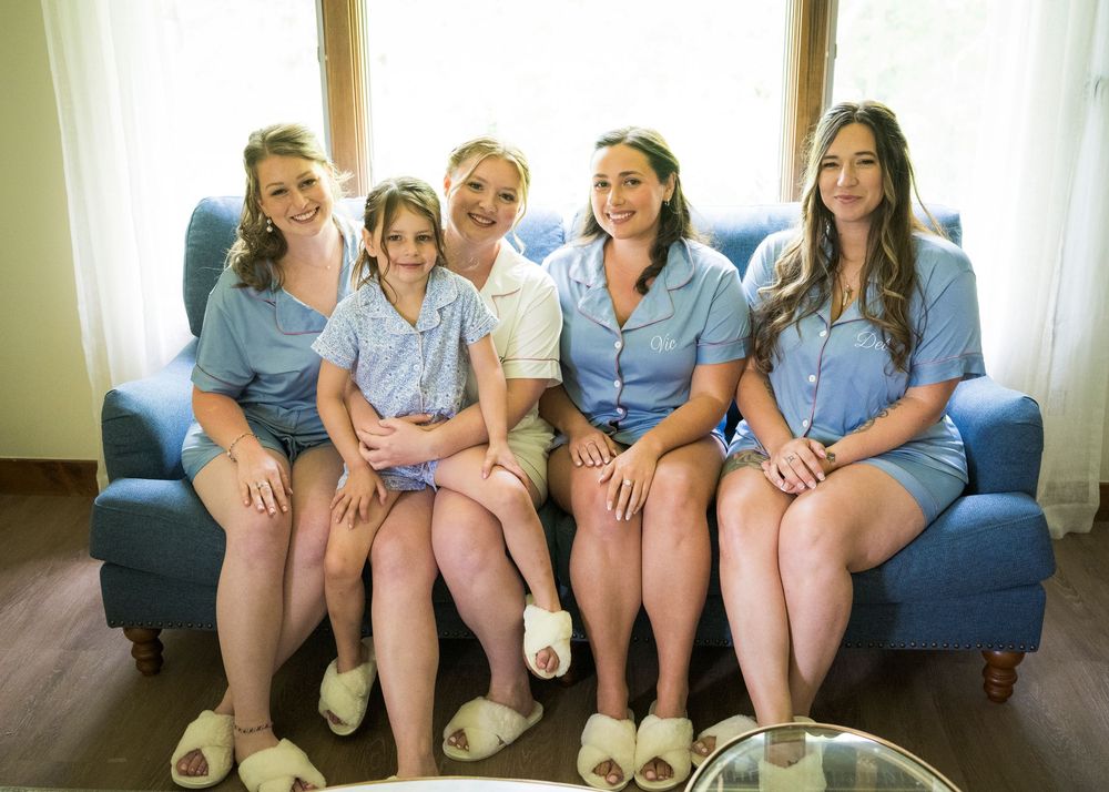 Five women in matching blue pajamas and white slippers sit together smiling on a blue armchair during bridal preparations.