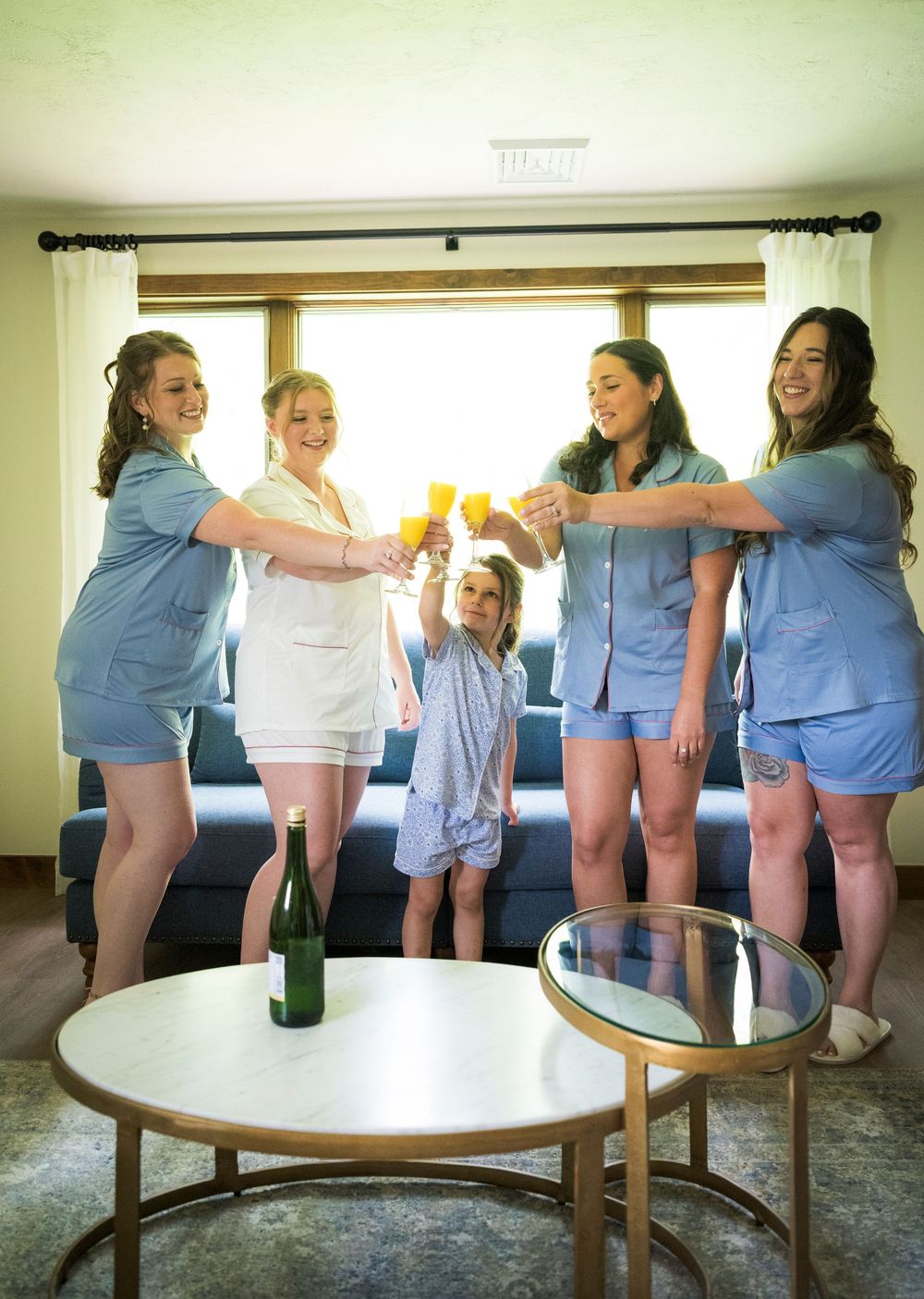 Five women in blue pajamas toast with champagne glasses beside a bottle on a round table in a bright living room.
