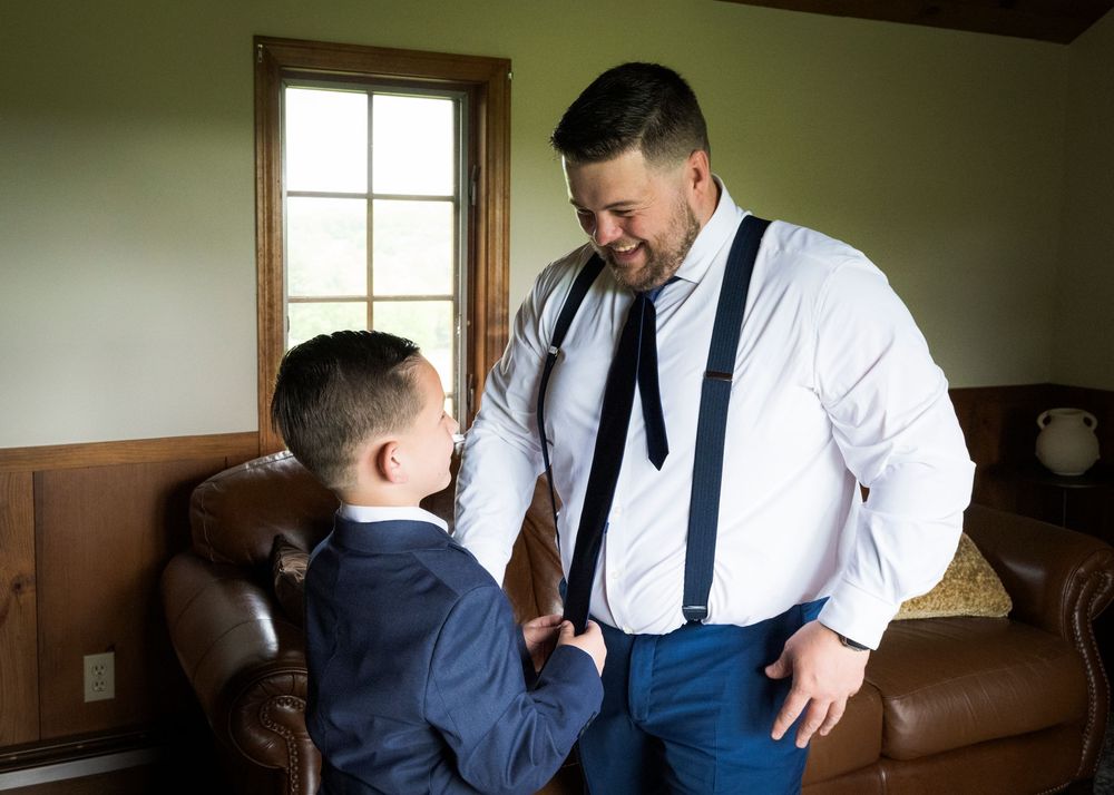 Groom in white shirt and suspenders shares a sweet moment with young boy in suit before wedding ceremony.