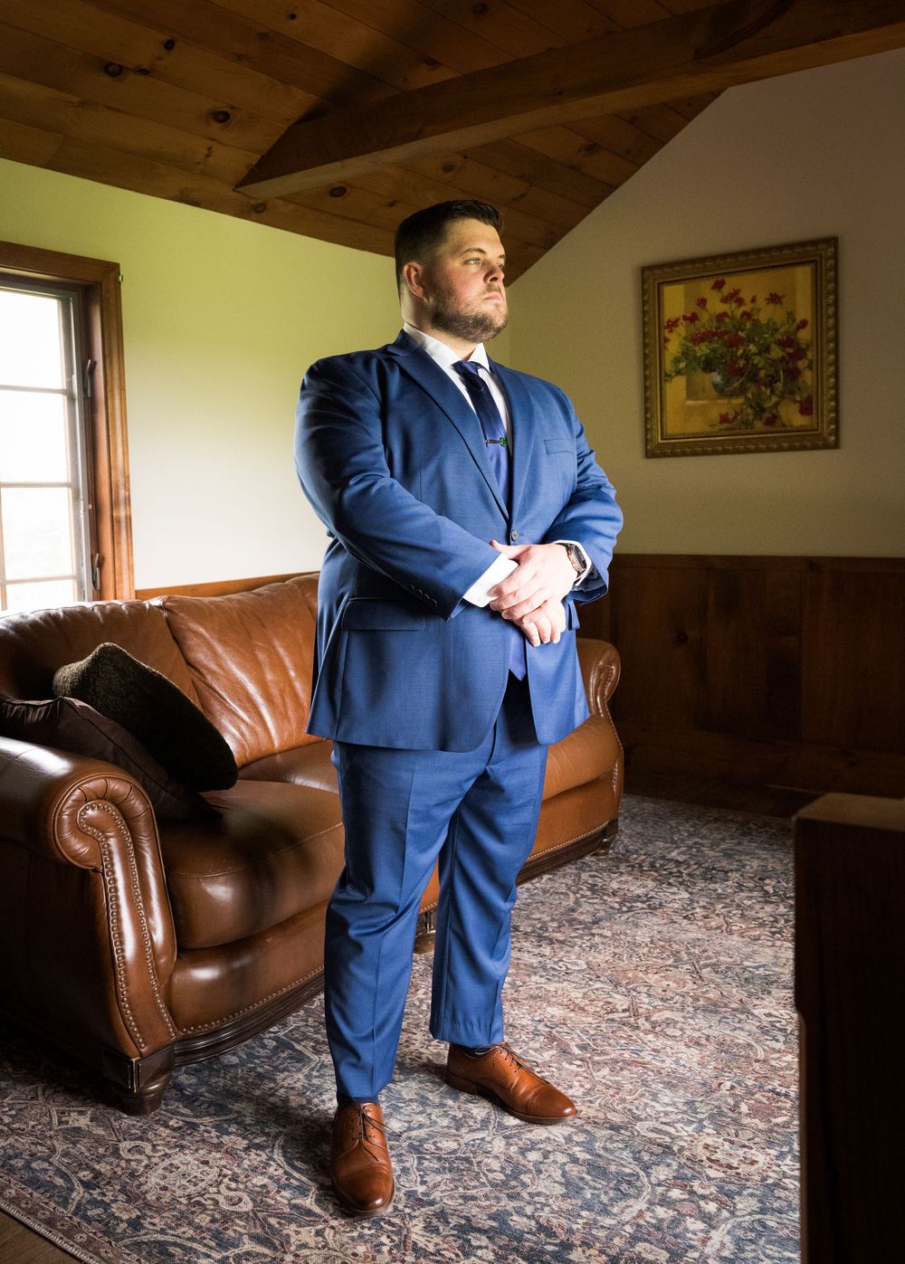 A groom in a blue three-piece suit with brown shoes stands confidently in a rustic room with a leather couch.