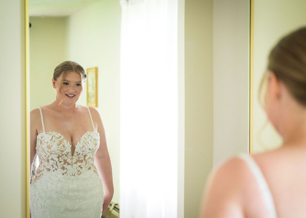 Bride in lace wedding gown smiling at her reflection in a full-length mirror before her wedding ceremony.