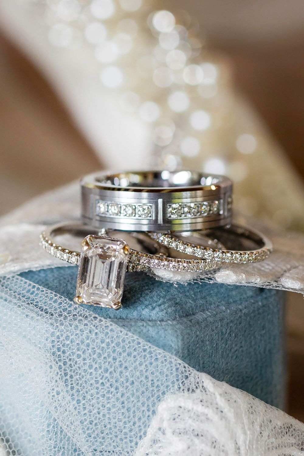 Elegant wedding ring set featuring emerald cut diamond engagement ring and matching bands on turquoise fabric with bokeh.