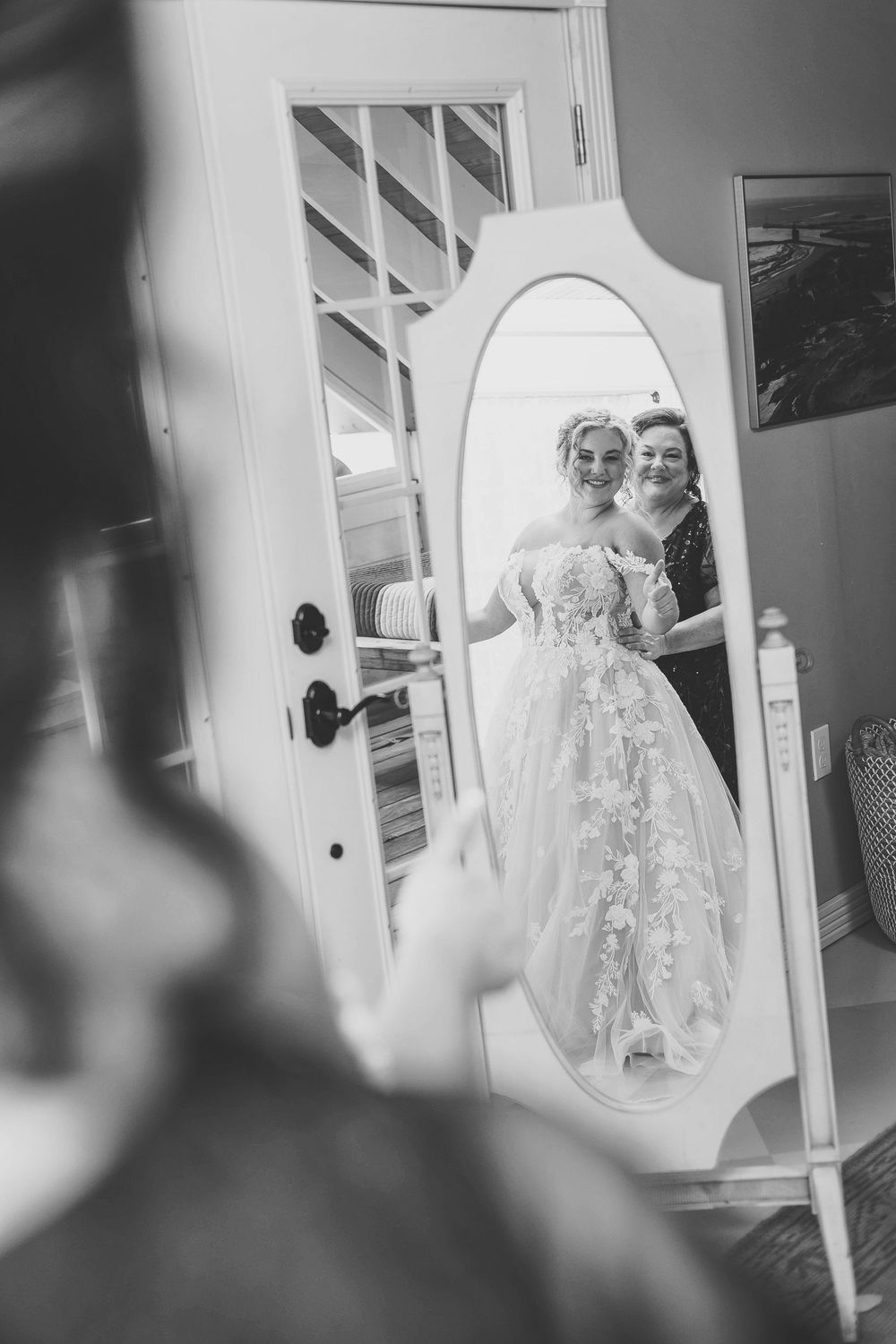 A black and white photo showing a bride in a lace wedding gown preparing in a reflection of a mirror while her mom zips her dress