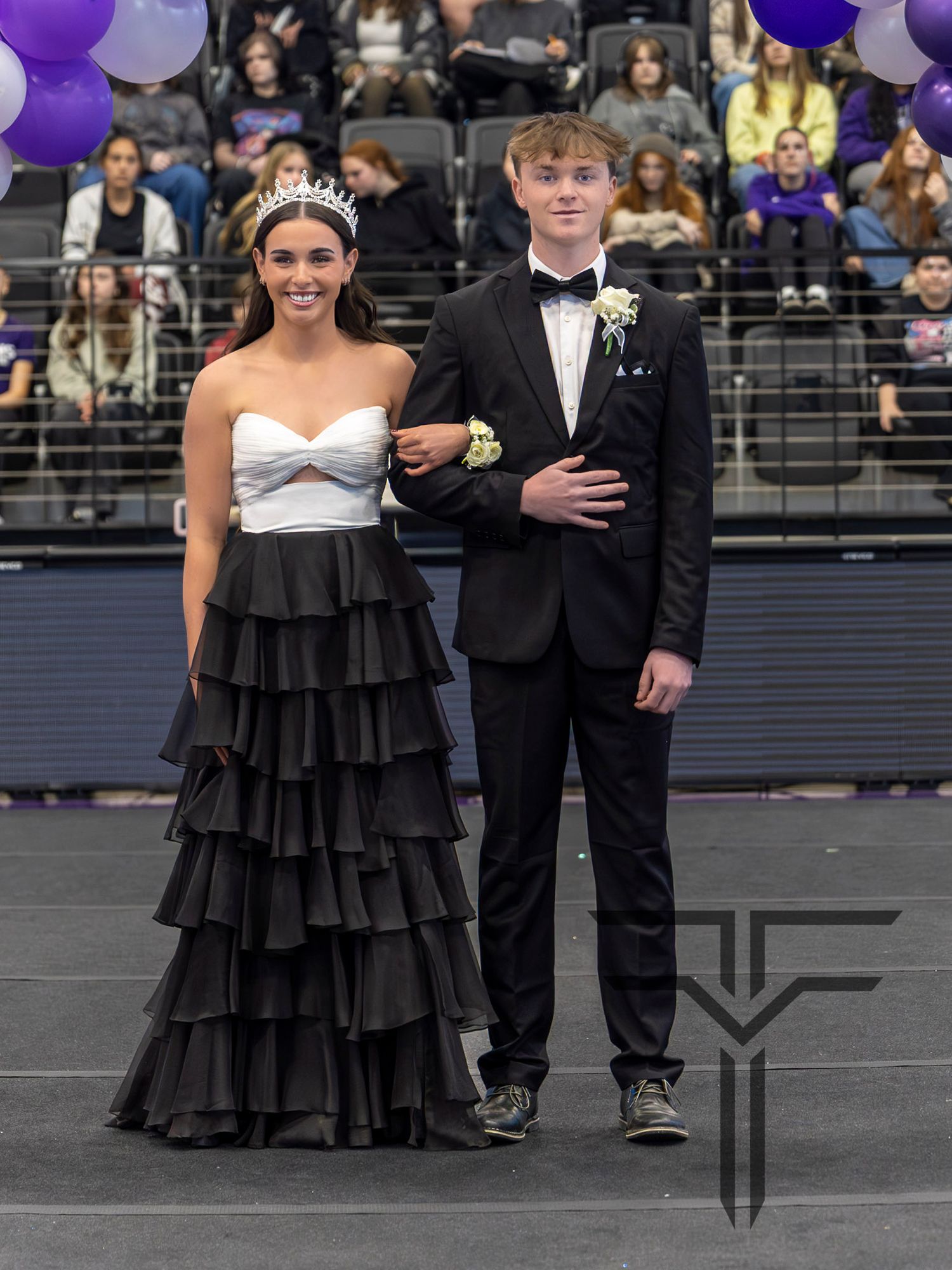 Teen couple in formal attire at homecoming, girl in black ruffled gown with tiara, boy in black tuxedo with boutonniere.