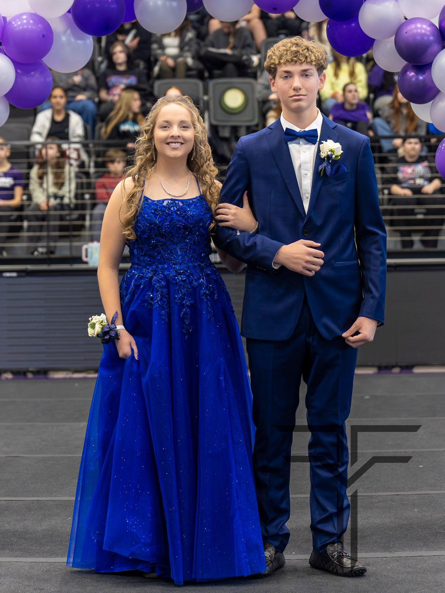 A young couple dressed in royal blue formal wear poses at a homecoming or prom event with purple and white balloons.