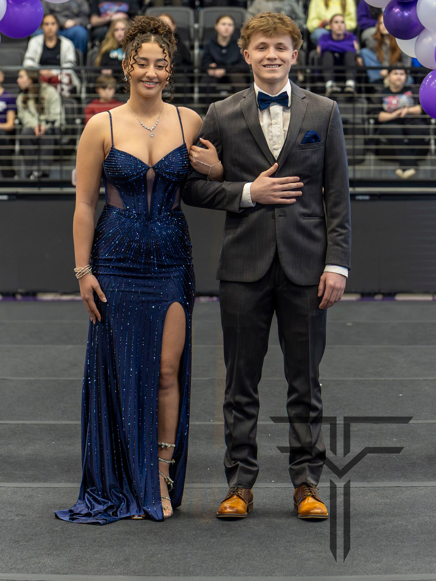 A couple dressed in formal attire poses at Color's Day in Elkins AR, wearing a blue sequin gown and gray tuxedo with bow tie.