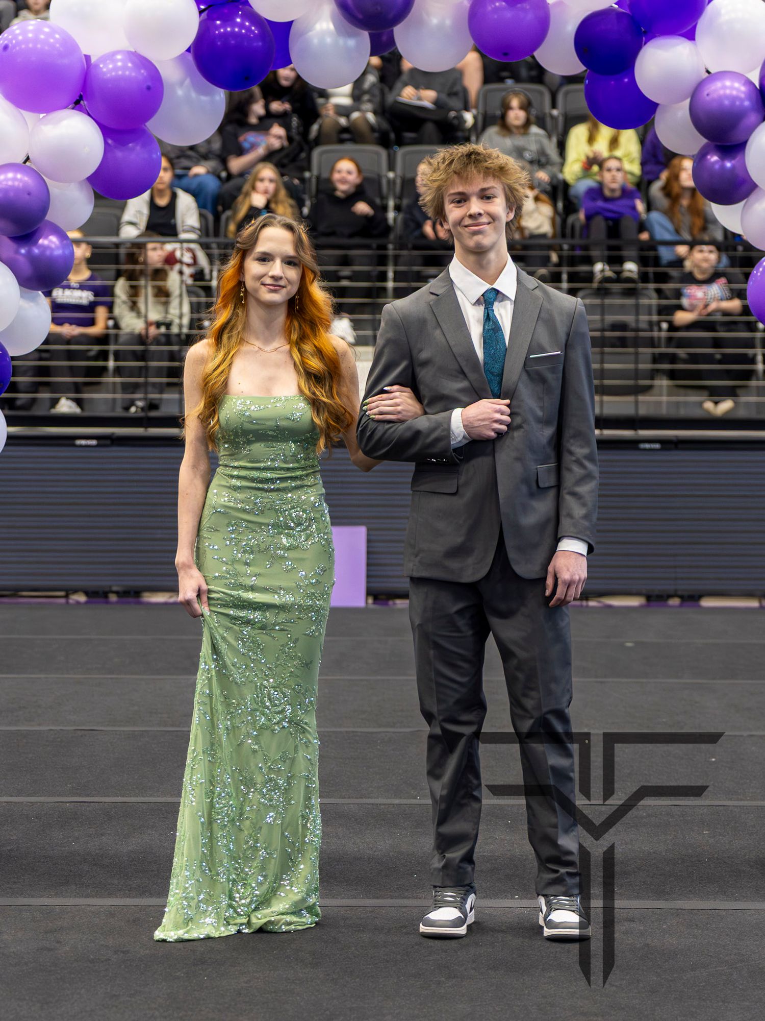 A young couple dressed in formal attire poses at prom, with purple and white balloons decorating the venue behind them.