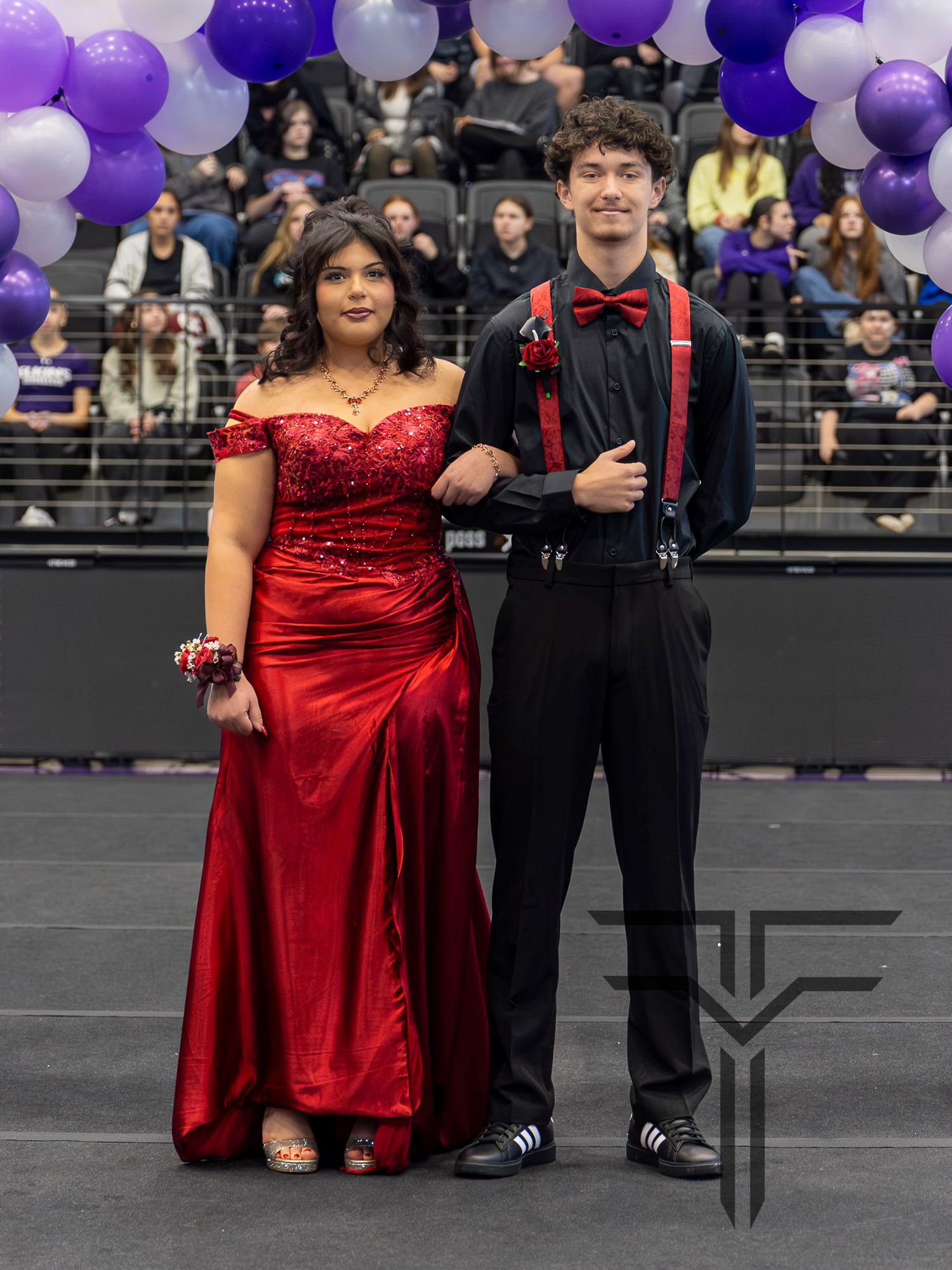 A couple in formal attire poses at prom, she in a red gown, he in black suit with red bow tie and suspenders.