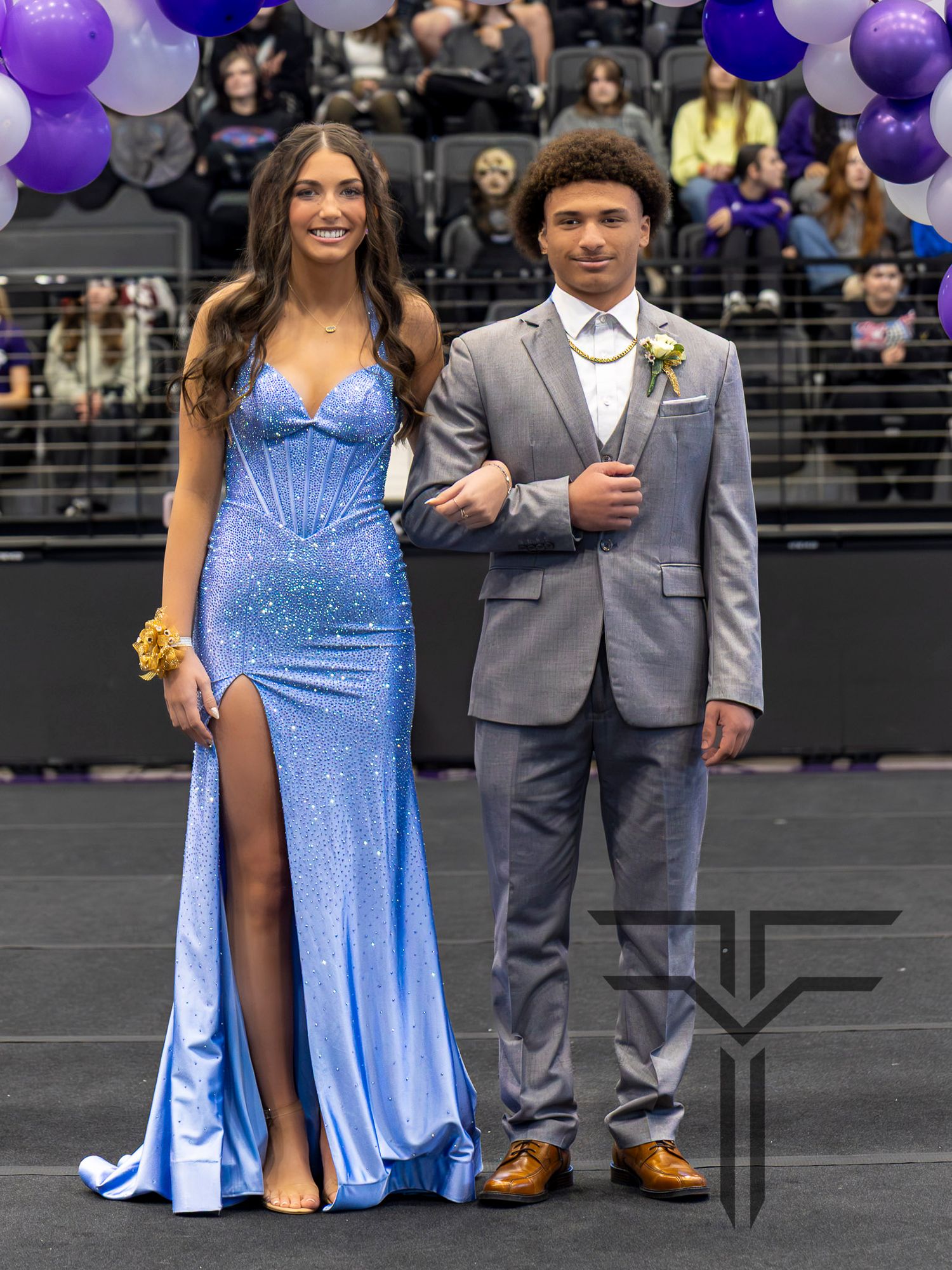 A couple dressed in prom attire, woman in blue sequin gown and man in gray suit, posing at a formal event.
