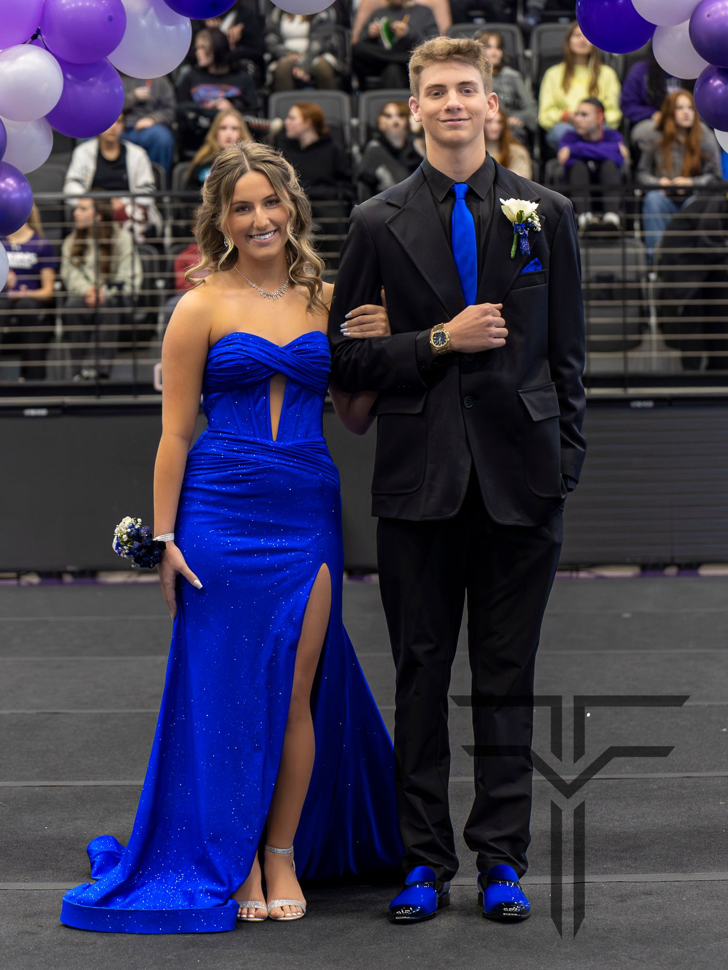A couple dressed in royal blue formal wear poses at a prom event in a decorated gymnasium.