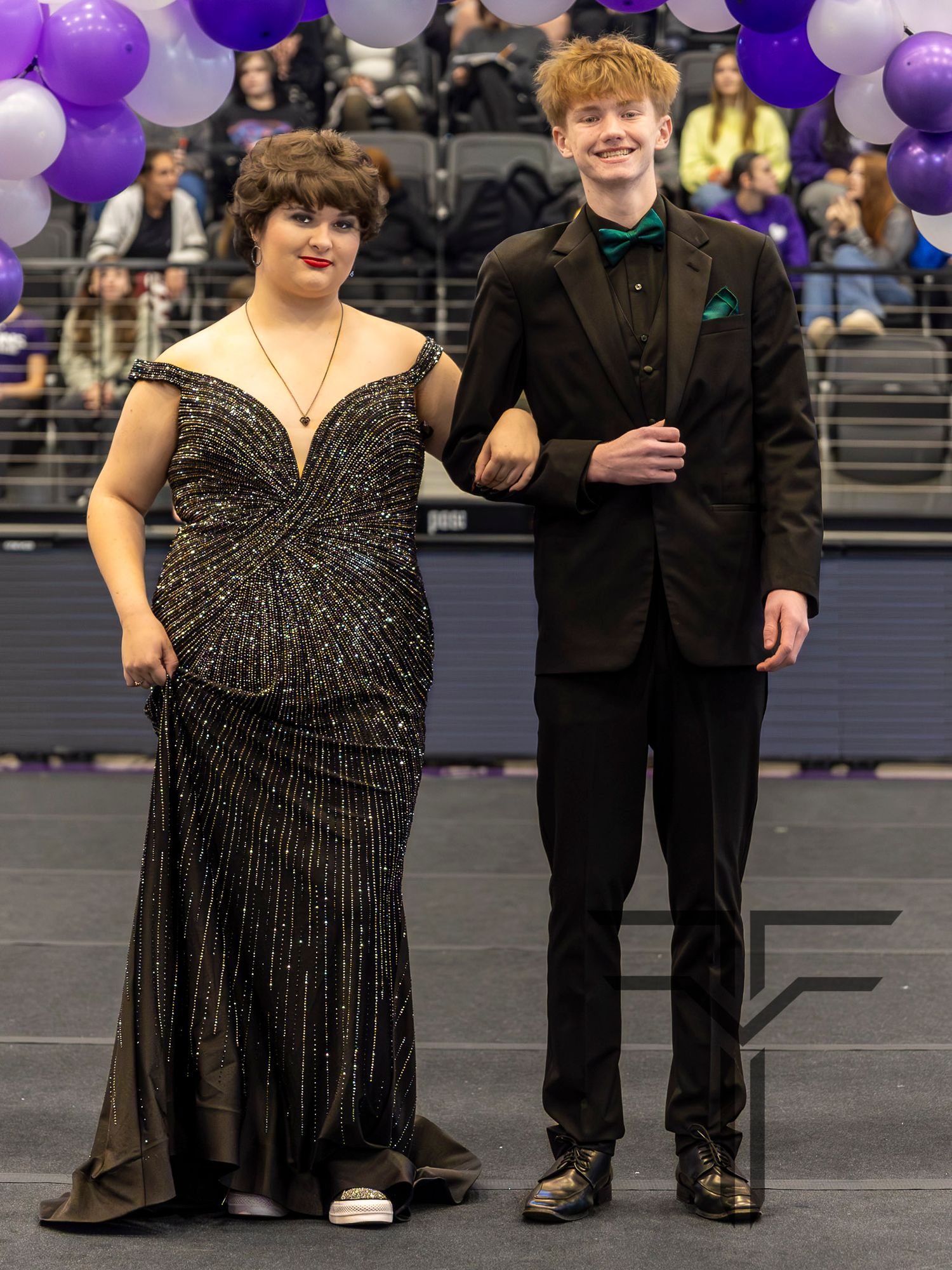 A young couple dressed in formal attire poses at a school dance, wearing a black gown and black tuxedo.