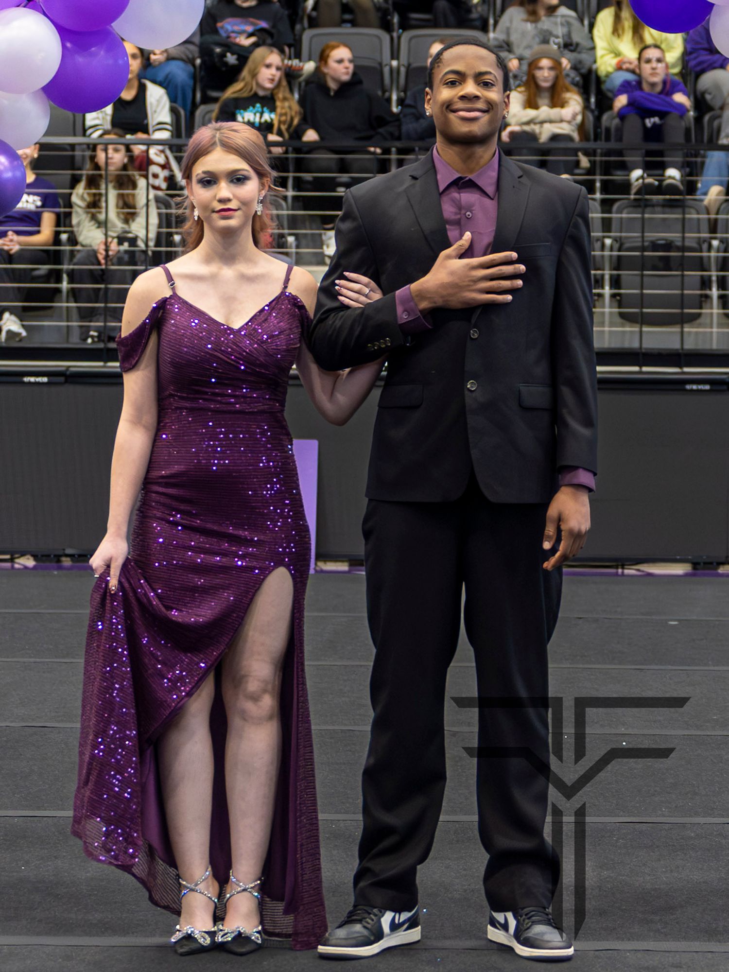 A young couple dressed in purple and black formal attire pose together at a school homecoming event.