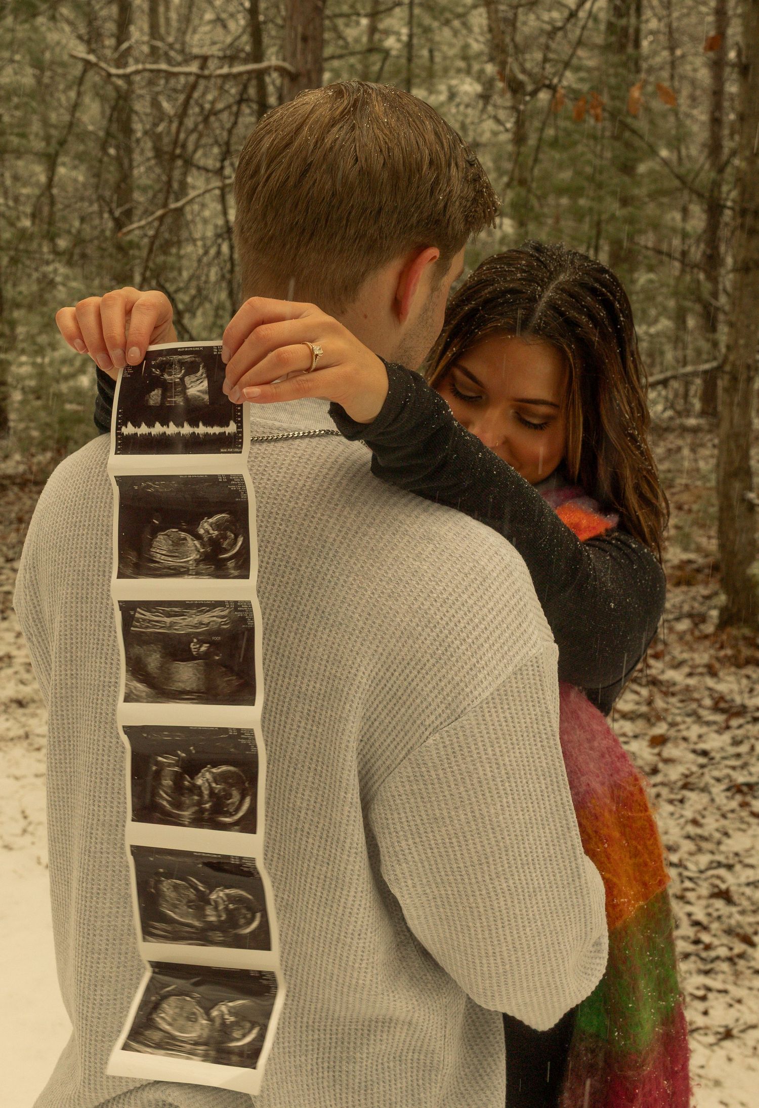 A couple shares an intimate moment while holding ultrasound photos in a wooded setting during winter. Creative Art By Bree LLC