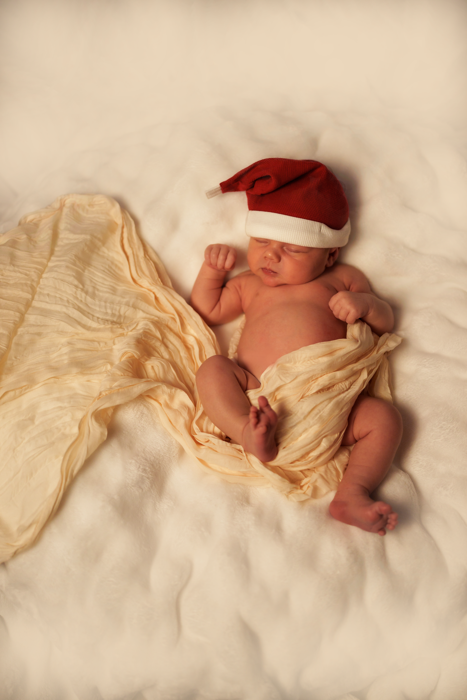 Baby lying on white bedding next to orange carrot prop in natural light setting.
