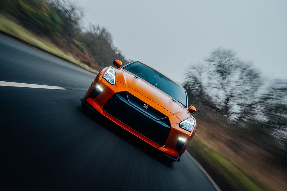 An orange Porsche GT3 RS sports car races along a winding road through scenic countryside.