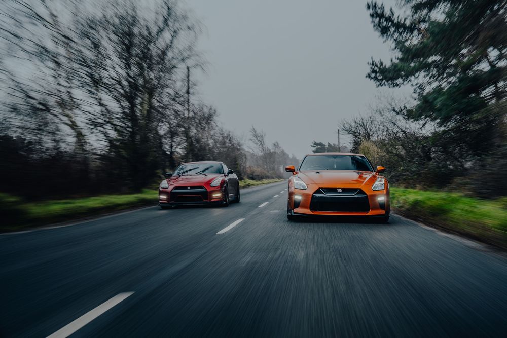 Orange sports car leads a pack of performance vehicles through a misty mountain road during an overcast day.