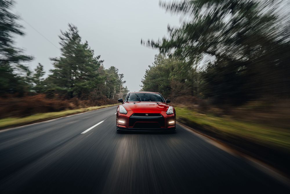 Red sports car speeding through a tree-lined road with motion blur effect in dramatic lighting conditions.