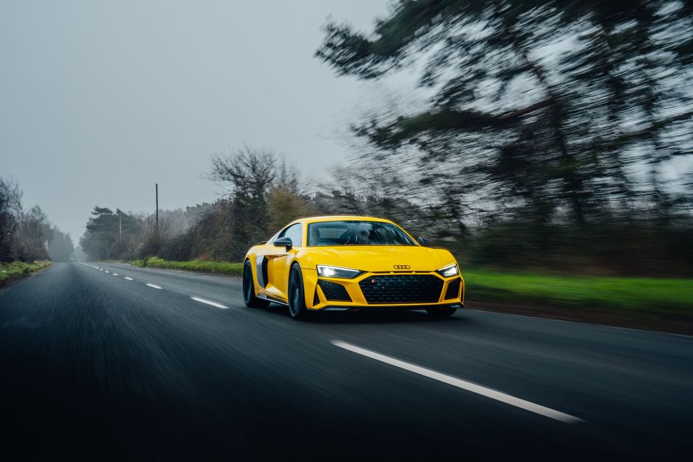 A yellow Audi R8 sports car drives on a winding road through misty green countryside.
