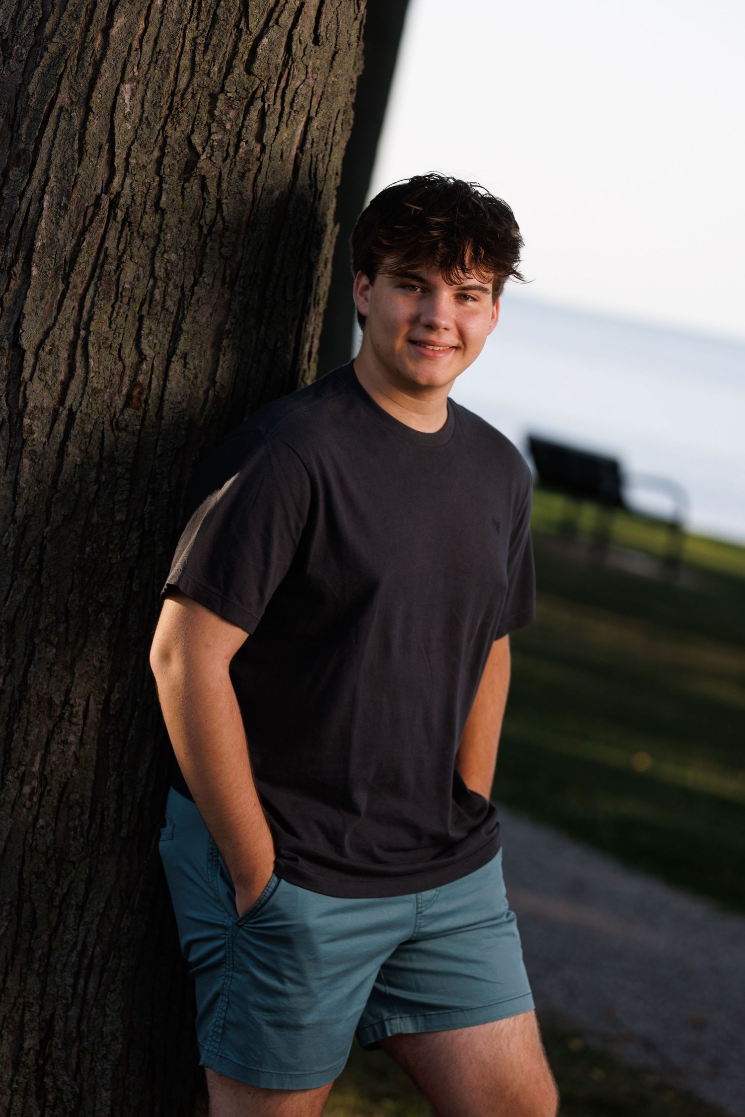 high school senior boy posed leaning against a tree in park setting.