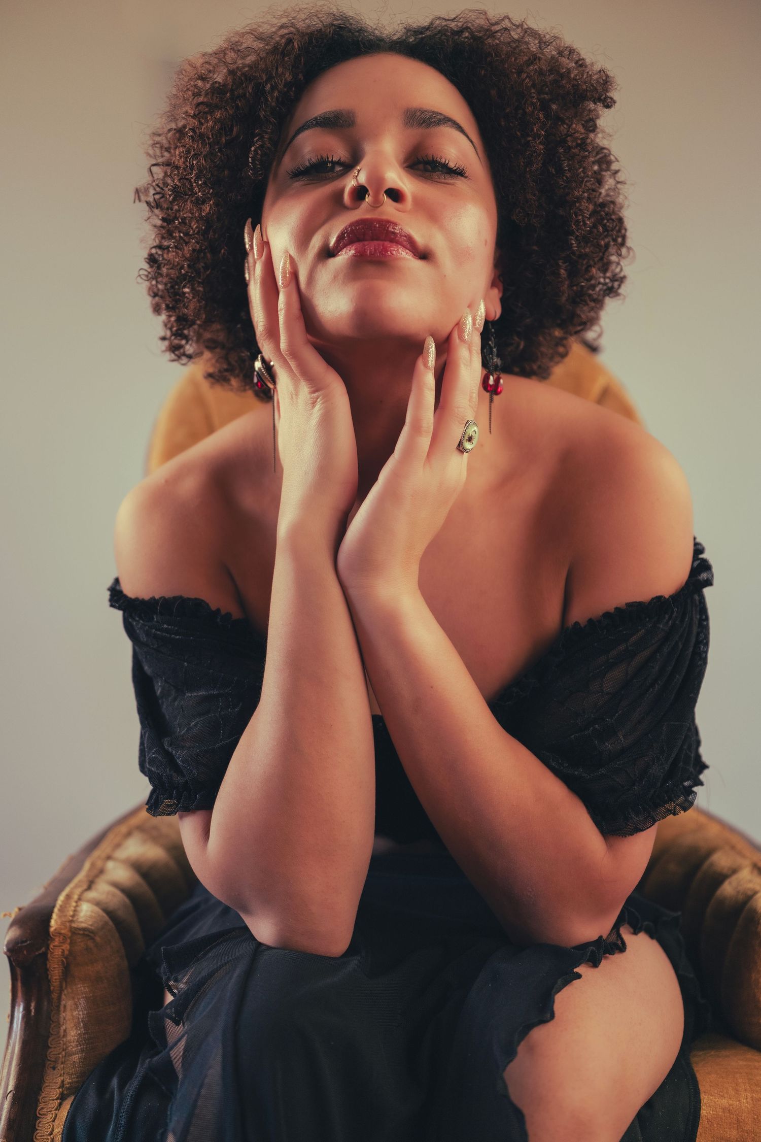 A glamour portrait features a model in a black off-shoulder top posing with hands touching face in a leather chair.