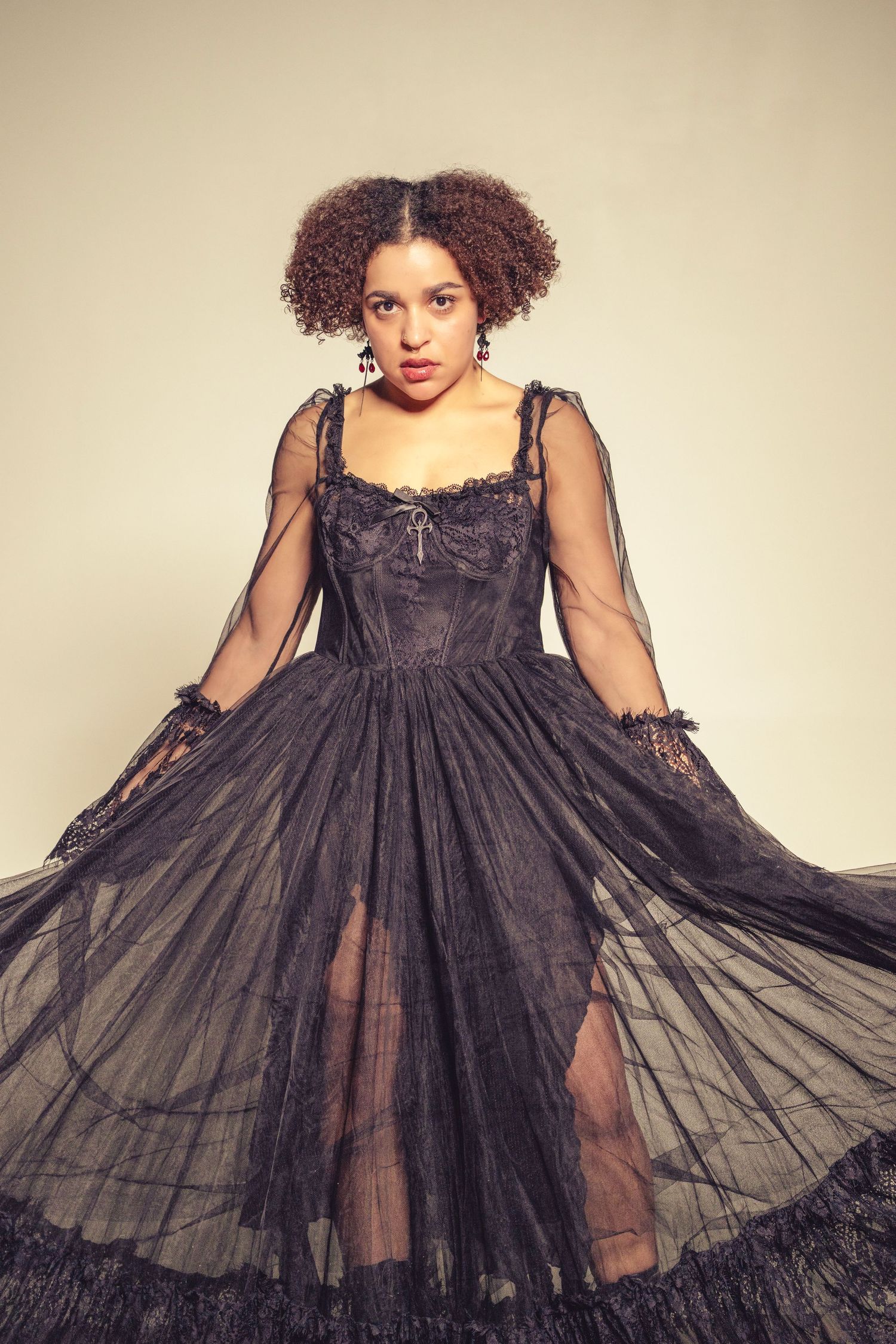 A dramatic fashion photo of a model in a flowing black sheer gown with lace details against a neutral backdrop.
