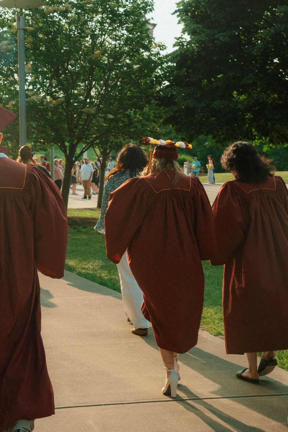 Students in maroon graduation gowns walking along a pathway surrounded by green trees.