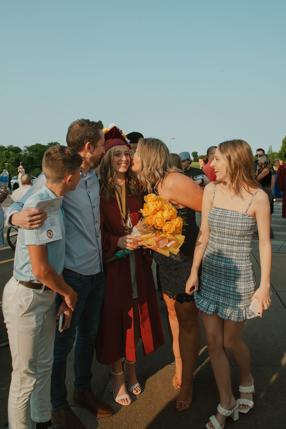 Group of graduates celebrating together outdoors during sunset with hugs and yellow flower bouquets.