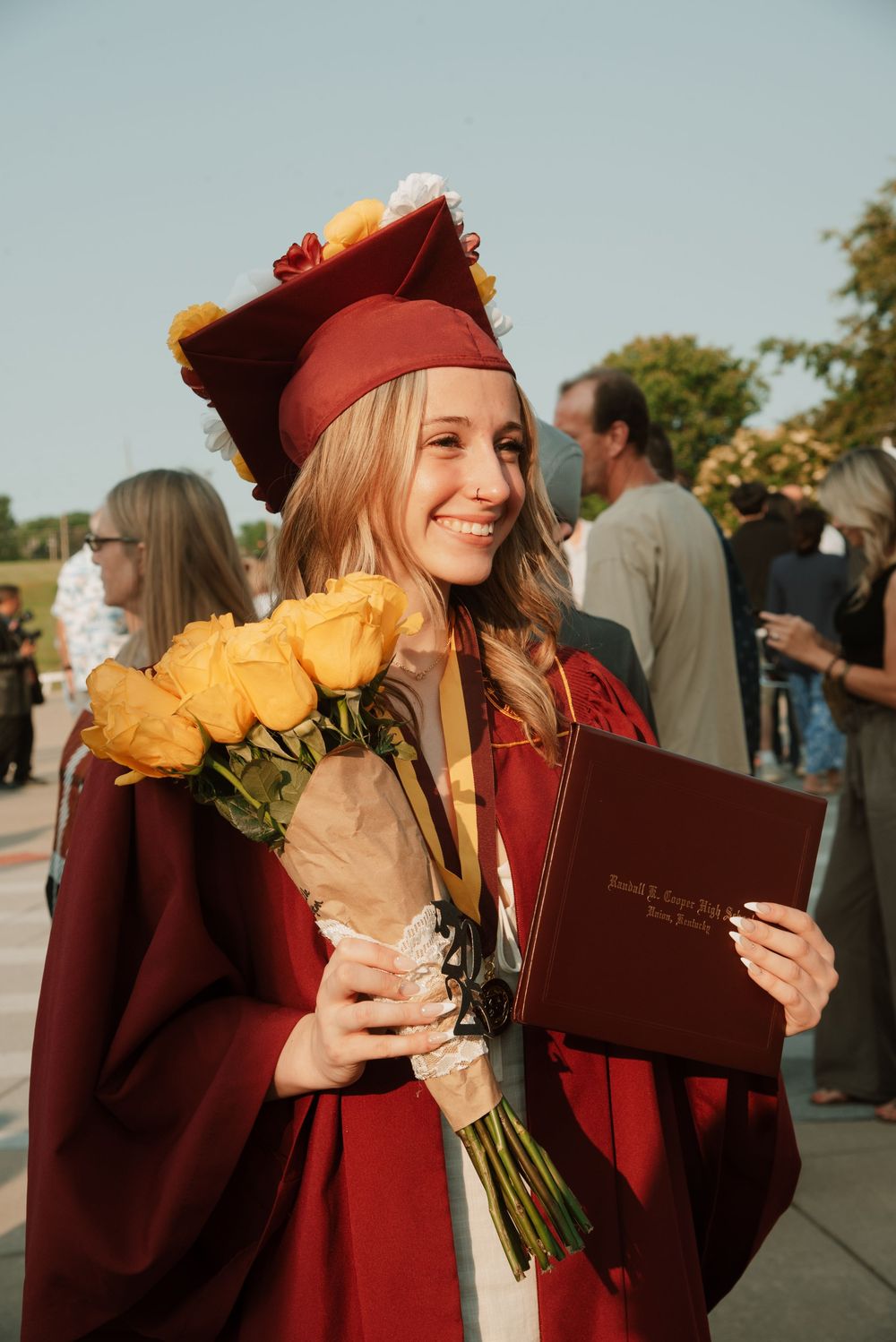 Graduate in maroon cap and gown holding yellow flowers and diploma during outdoor ceremony.