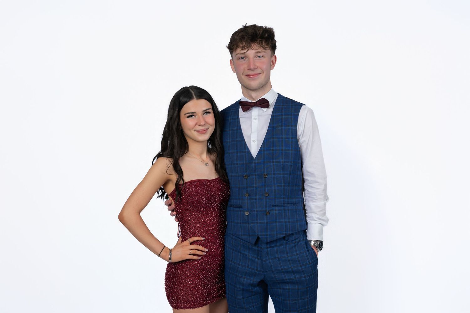A couple in formal attire poses together, with her wearing a burgundy dress and him in a blue vest and white shirt.