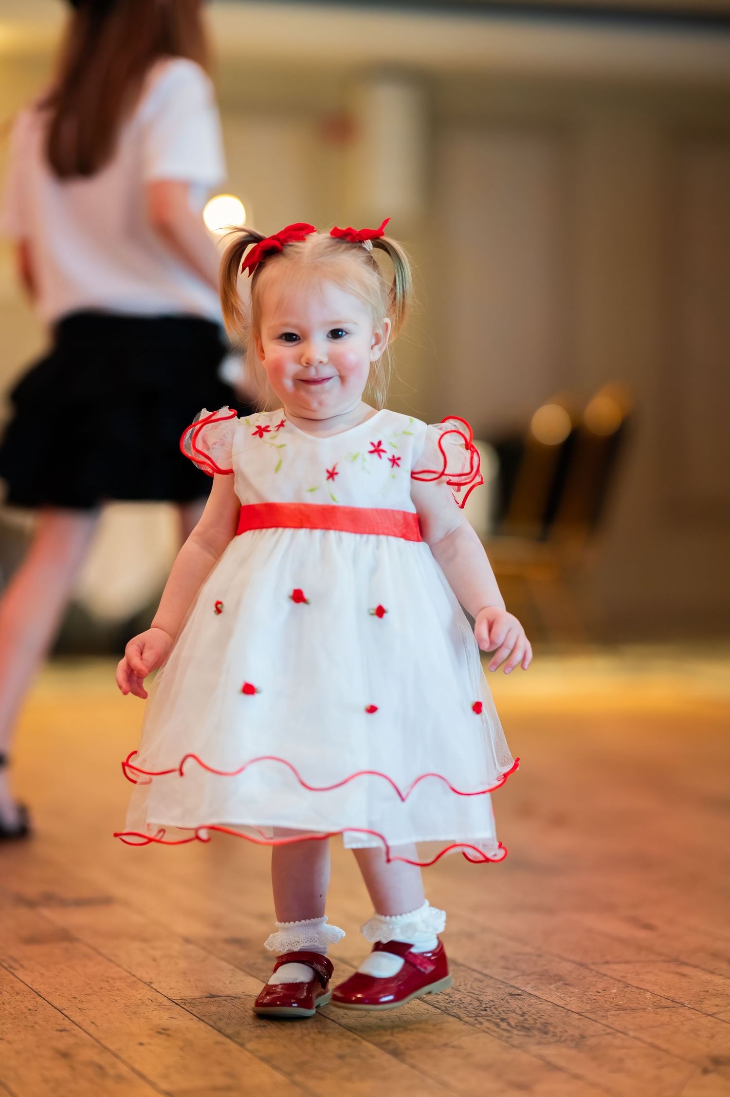 A small child in a white dress with red trim and bows twirls on a hardwood floor.
