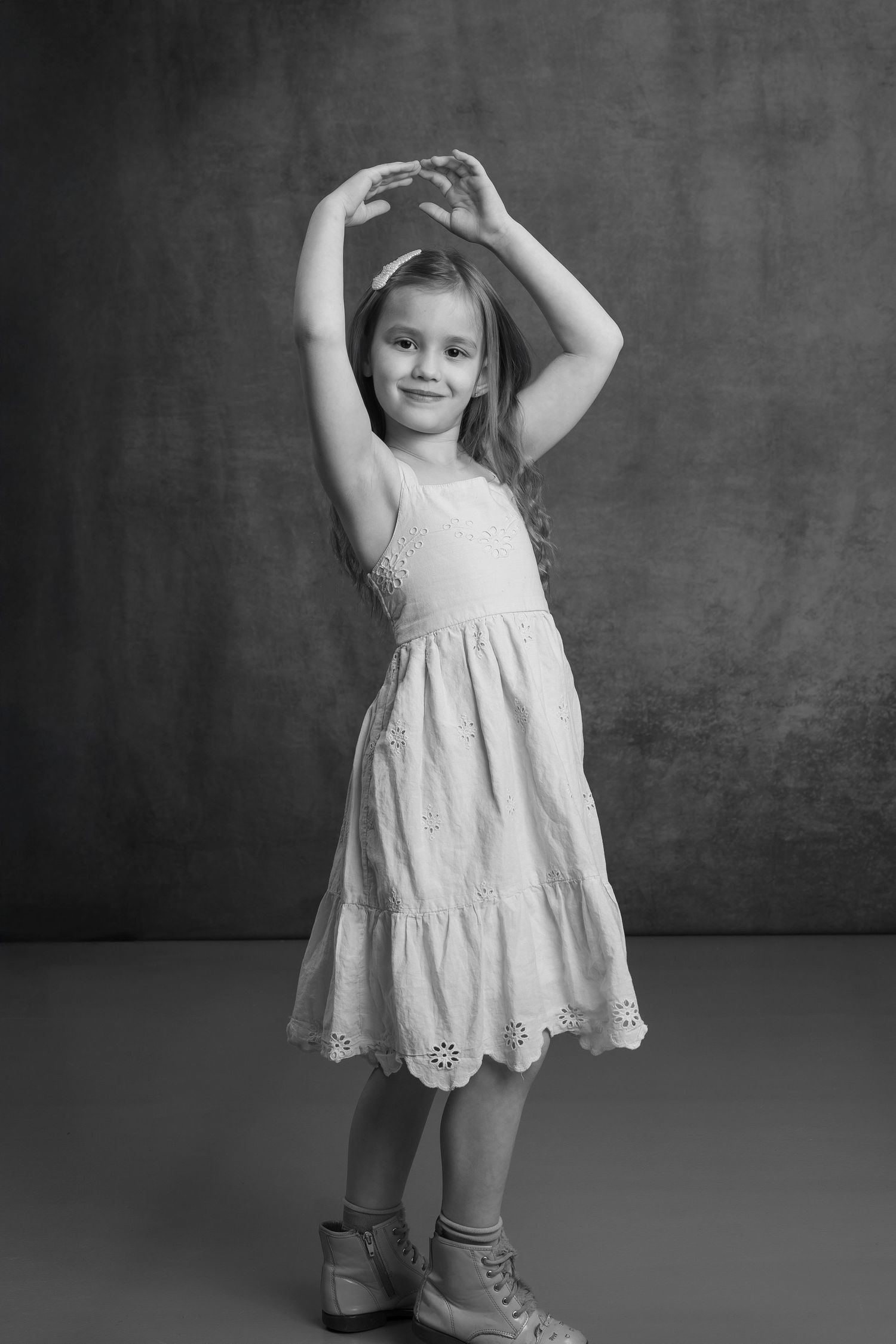 A young girl in a white eyelet dress poses gracefully with arms raised overhead in a black and white studio portrait.