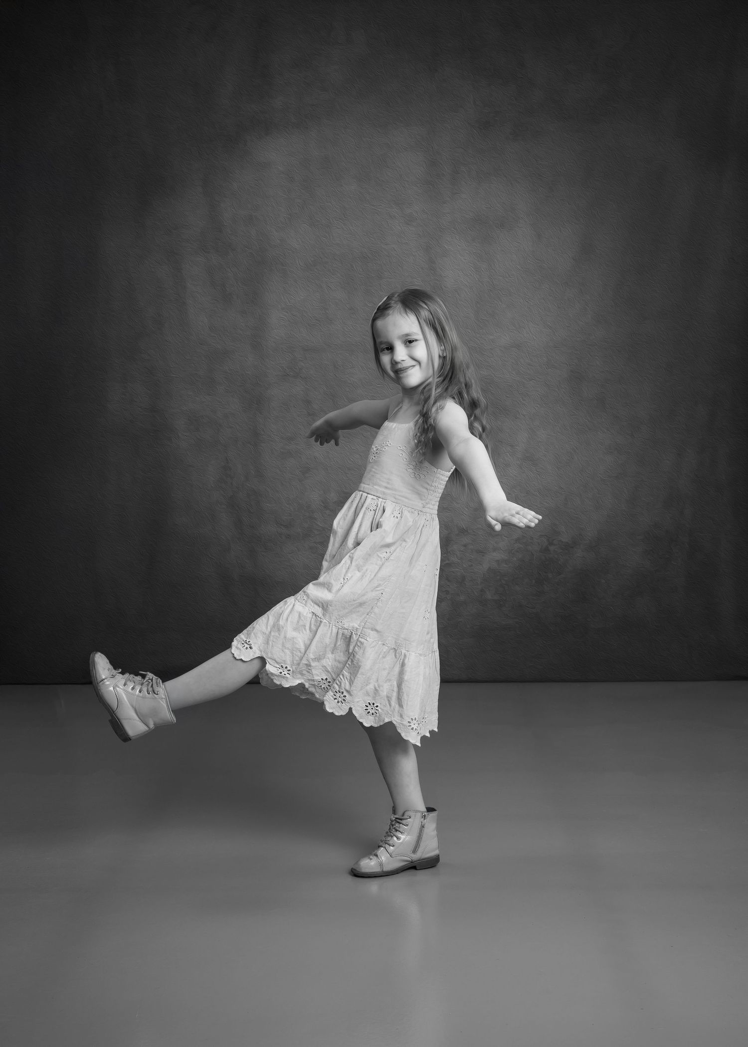 A cheerful young girl in a white eyelet dress kicks her leg up playfully in a black and white studio photo.