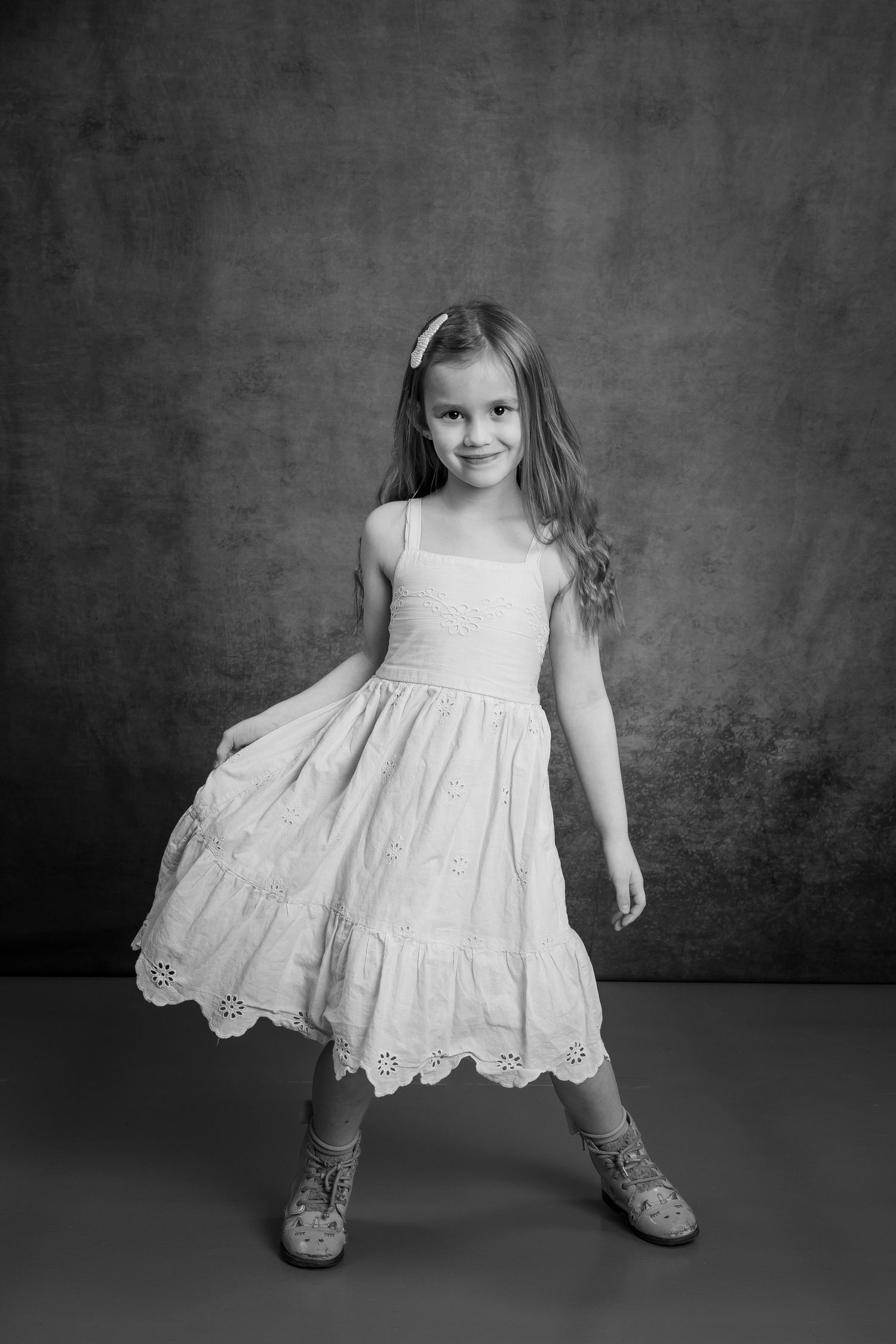 A smiling young girl in a white eyelet dress poses confidently in a classic black and white studio portrait.