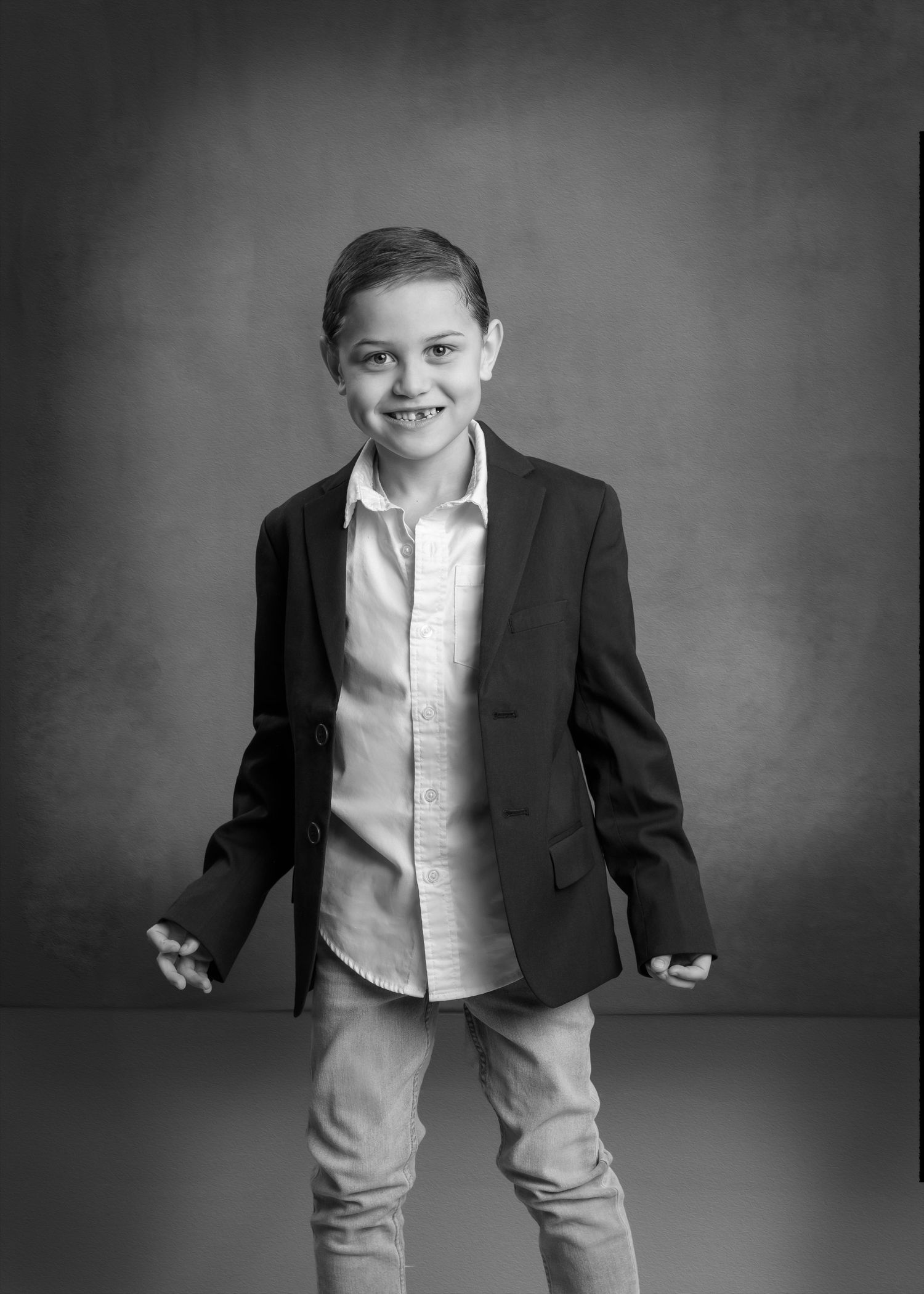 A young boy in a blazer and dress shirt smiles confidently in a stylish black and white studio portrait.