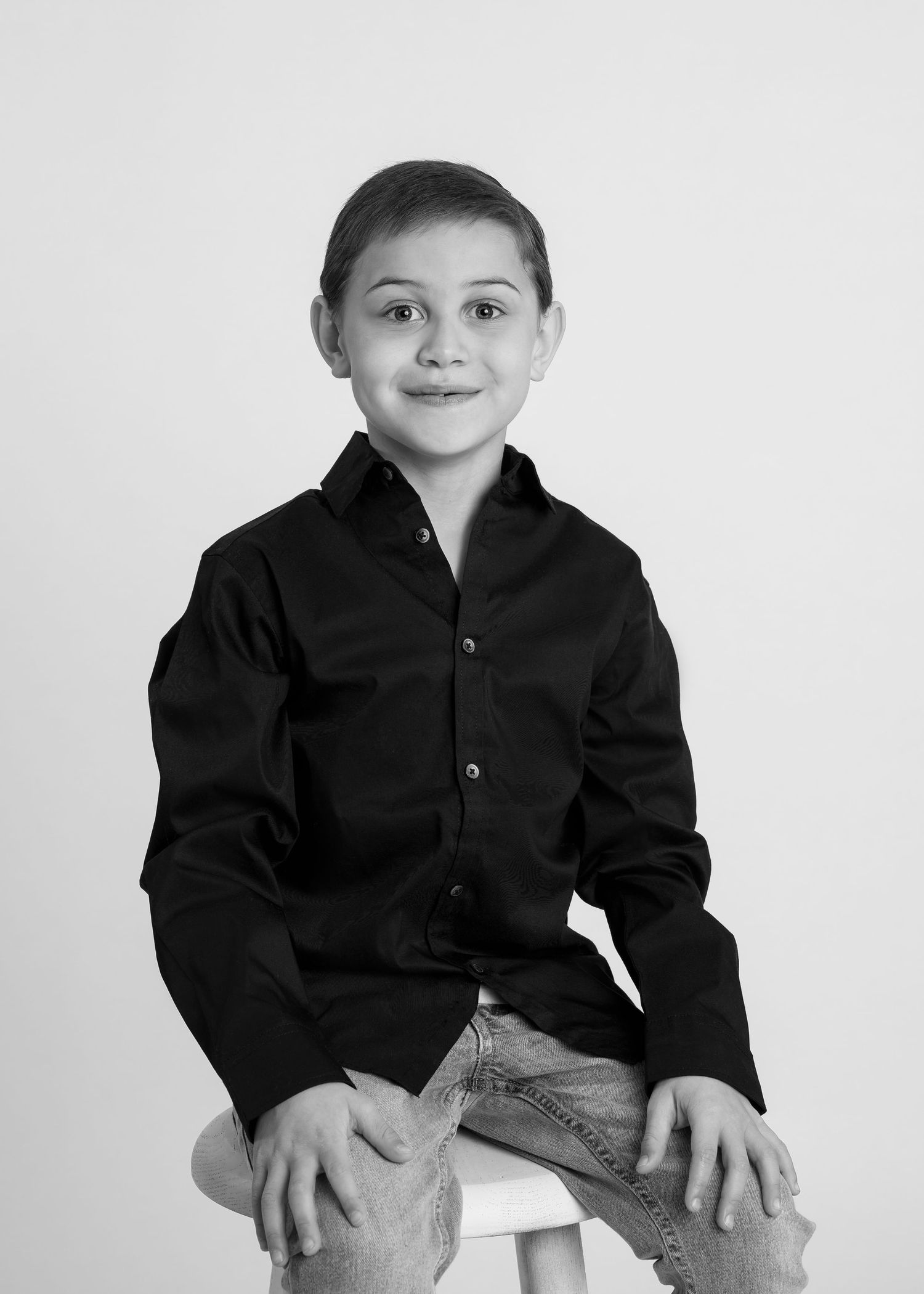 A young boy in a black button-up shirt sits on a stool smiling in a clean black and white studio portrait.