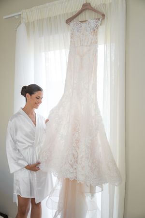 A lace wedding dress hangs by a window while a woman in a white robe admires it.